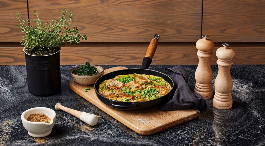 Gusseisenpfanne von Le Creuset mit Pfannengericht mit Fleisch und Gemüse, dekoriert mit Kräutern, auf Holzbrett. Kräutertopf, Gewürzschälchen und Pfeffermühlen daneben.