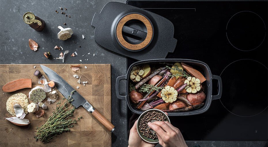 Oberansicht von Zutaten und einer Kochstelle, eine Hand fügt Gewürze zu einem Bräter von Fiskars mit Gemüse auf einem Herd hinzu.