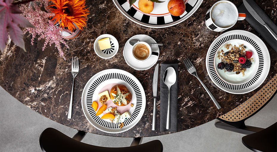 Ein mit Geschirr der Memphis Collection von Villeroy &amp; Boch gedeckter Frühstückstisch mit Marmortafel; Teller mit Obst, Joghurt, Besteck, Espresso, dekorative Blumen und Servietten.