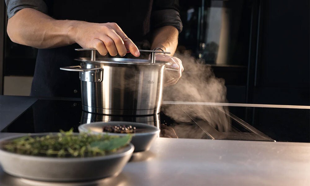 Person kocht an einem Induktionsherd, hebt den Deckel eines dampfenden Edelstahltopfs von Fissler. Kräuter in Schalen im Vordergrund.