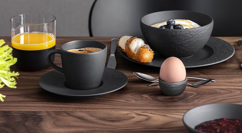 Frühstücksszene mit Kaffee, Orangensaft, Eierbecher von Villeroy &amp; Boch, Joghurt mit Heidelbeeren, Gebäck und Besteck auf Holztisch.