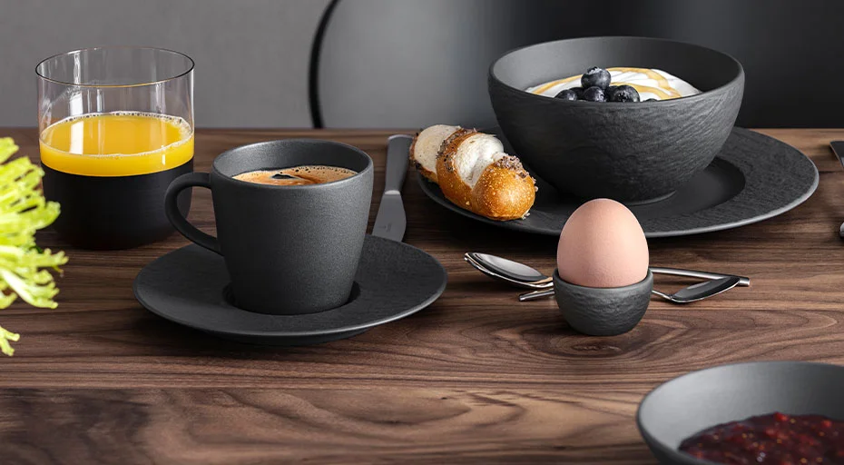 Frühstücksszene mit Kaffee, Orangensaft, Eierbecher von Villeroy &amp; Boch, Joghurt mit Heidelbeeren, Gebäck und Besteck auf Holztisch.