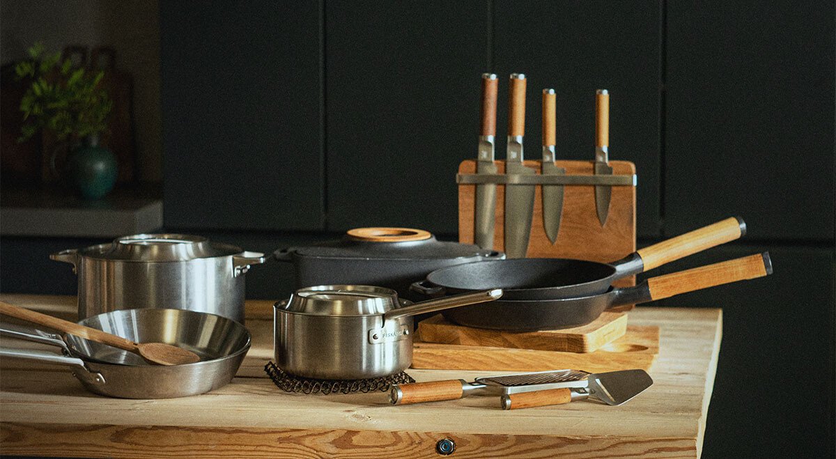 Verschiedene Pfannen, Töpfe und Bräter von Fiskars auf Holz-Arbeitsplatte neben Messerblock.