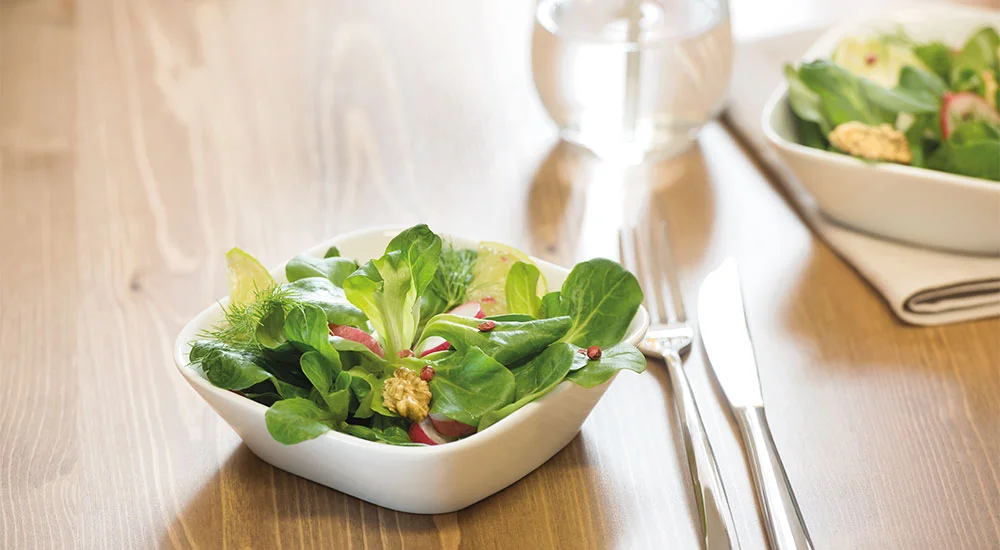 Salatschale von Villeroy &amp; Boch mit frischen Blättern, Radieschen und Walnüssen auf Holztisch neben Besteck und Wasserglas.