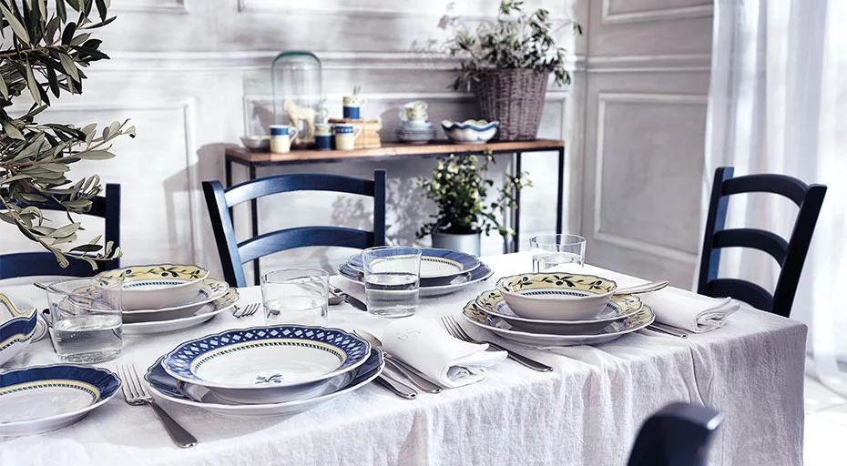 Elegant gedeckter Tisch mit Porzellangeschirr von Hutschenreuther, Gläsern und Besteck auf weißer Tischdecke, blaue Stühle im Hintergrund.
