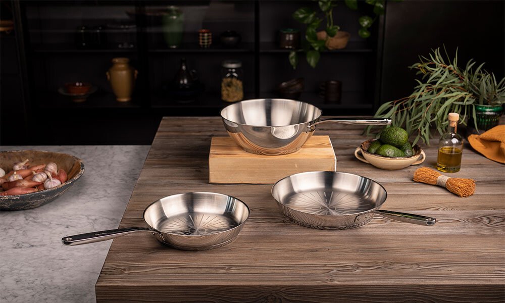Edelstahlpfannen von Fiskars auf Holztisch, dekoriert mit Kräutern und Gemüse im Hintergrund, in moderner Küche.