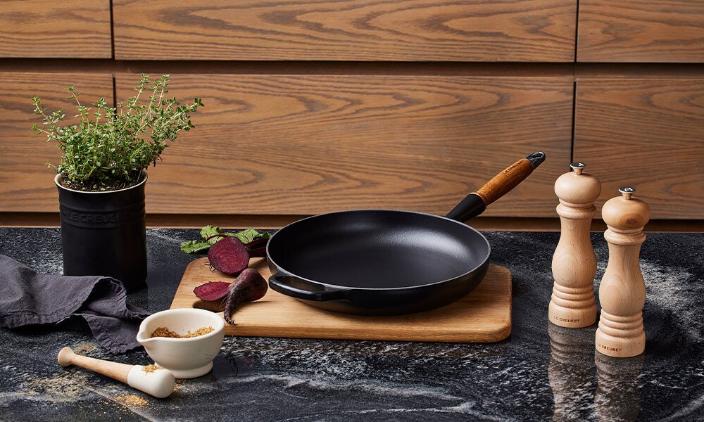 Bratpfanne von Le Creuset auf Schneidebrett, daneben Pfeffermühlen, Kräutertopf, Mörser und rote Bete auf dunkler Küchenarbeitsplatte.