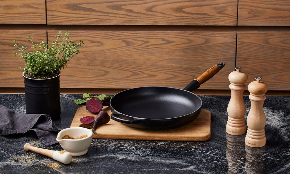 Bratpfanne von Le Creuset auf Schneidebrett, daneben Pfeffermühlen, Kräutertopf, Mörser und rote Bete auf dunkler Küchenarbeitsplatte.