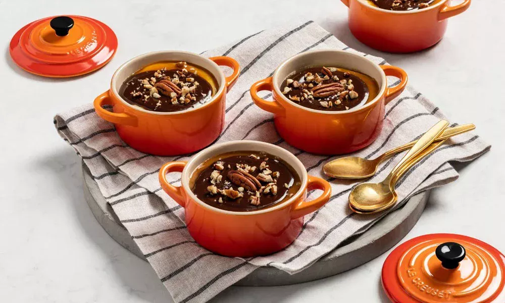 Vier orangene Le Creuset Mini-Cocotten mit Schokoladenpudding, bestreut mit gehackten Pekannüssen, auf einem gestreiften Handtuch.