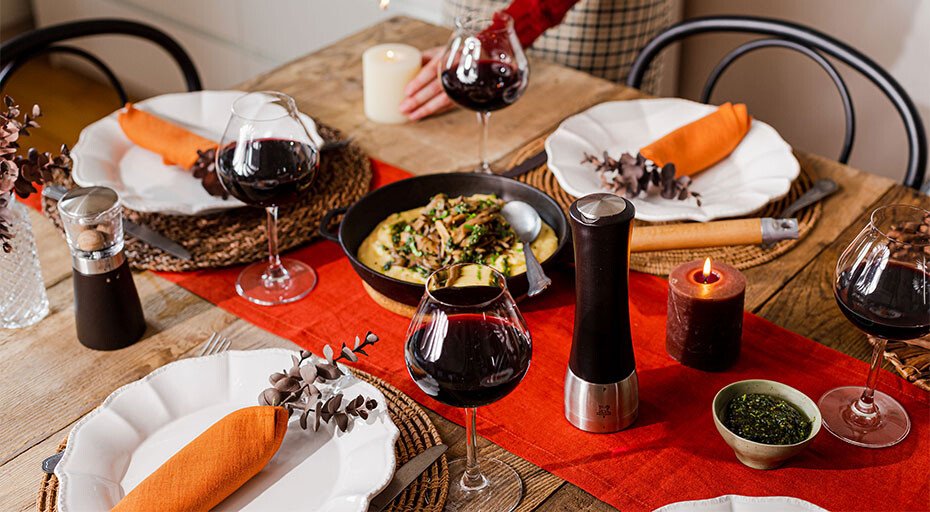Gedeckter Tisch mit Weingläsern, Teller, orange Servietten, Pfeffermühle und Kerzen auf rotem Tischläufer.