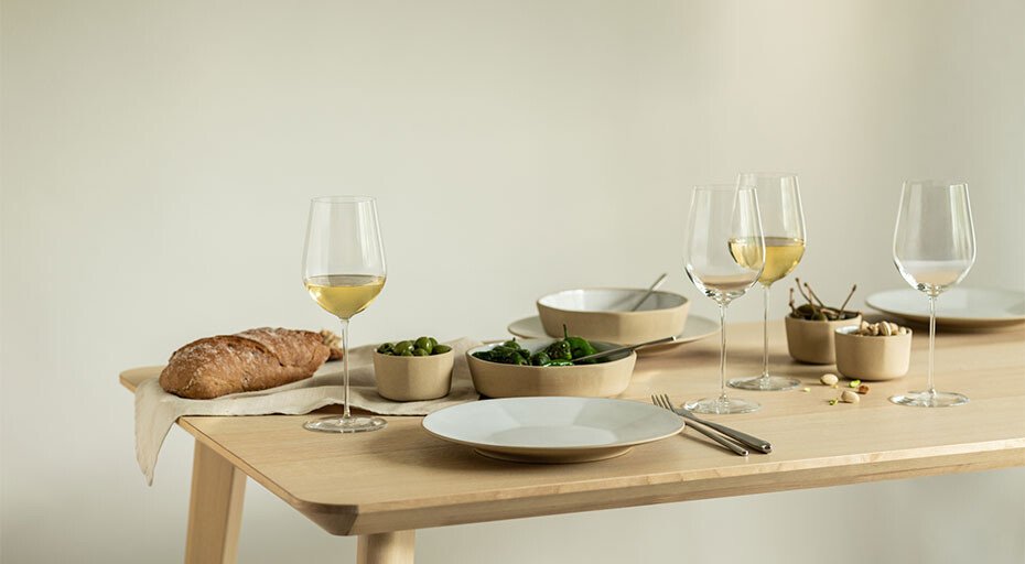 Gedeckter Tisch mit Weingläsern von Zwiesel Glas, Tellern, Schalen mit grünem Gemüse und Brot auf hellem Holztisch.