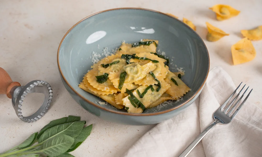 Schüssel mit Ravioli. Daneben ein Pastaschneider, Salbeiblätter und eine Gabel auf Stoffserviette.