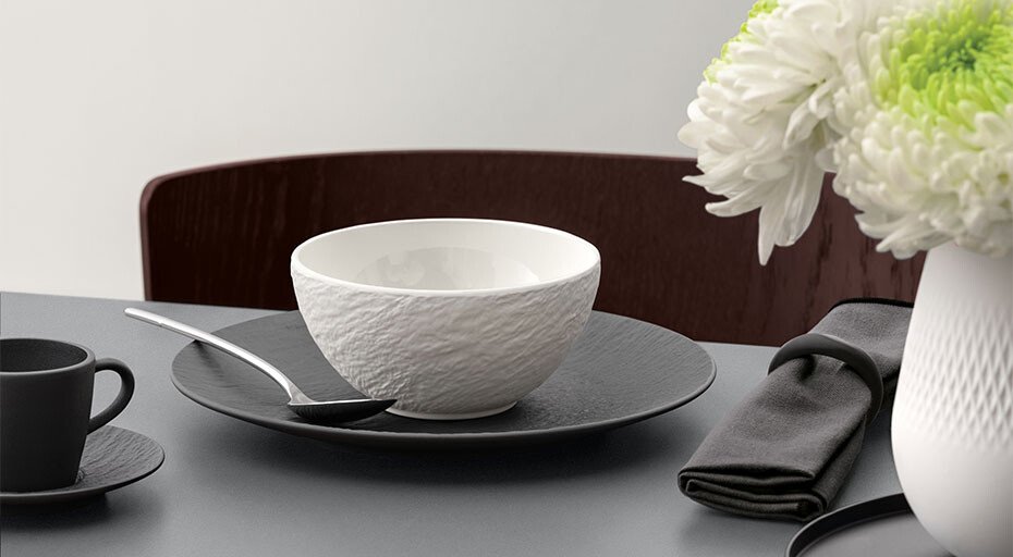 Tischgedeck mit Villeroy &amp; Boch Manufacture Ice Schale, Teller, Teetasse, Löffel, dunkler Serviette und Blumenvase mit weißen Chrysanthemen.