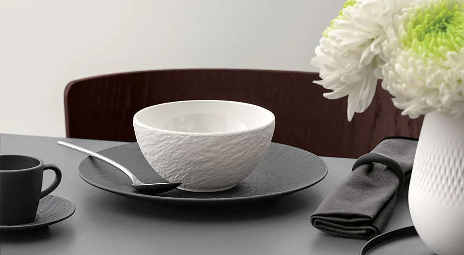 Tischgedeck mit Villeroy &amp; Boch Manufacture Ice Schale, Teller, Teetasse, Löffel, dunkler Serviette und Blumenvase mit weißen Chrysanthemen.