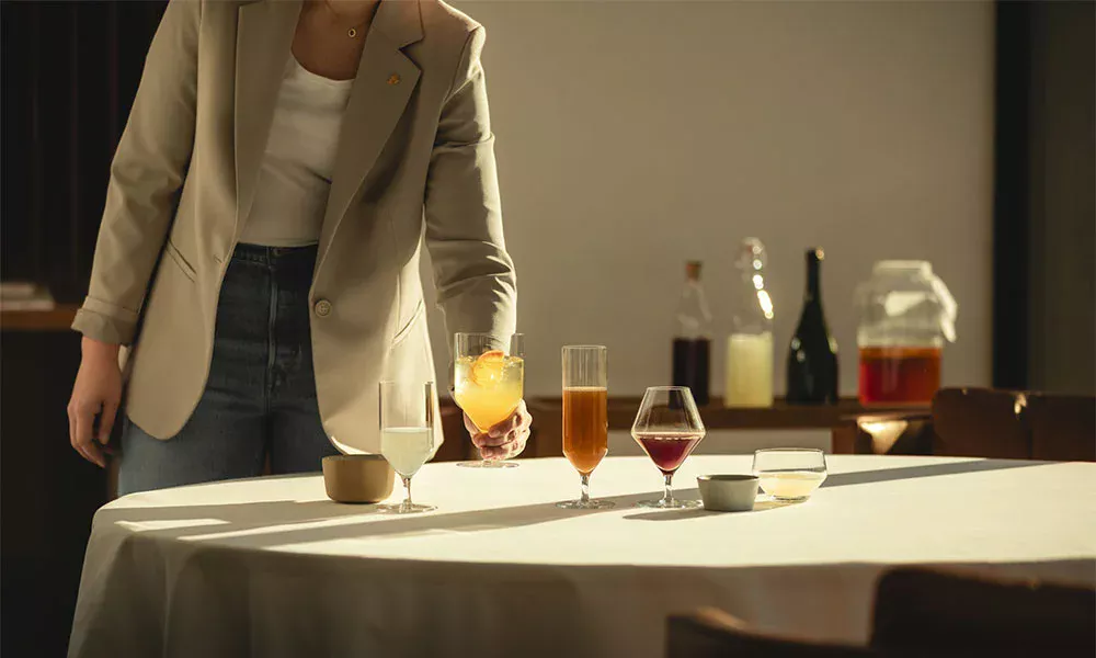 Person richtet Gläser der Serie Début von Zwiesel Glas mit Getränken auf einem Tisch her, im Hintergrund Flaschen und ein Gefäß. Stimmungsvolles Licht.