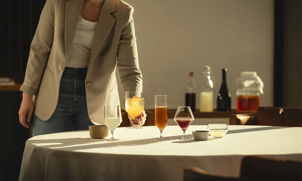 Person richtet Gläser der Serie Début von Zwiesel Glas mit Getränken auf einem Tisch her, im Hintergrund Flaschen und ein Gefäß. Stimmungsvolles Licht.
