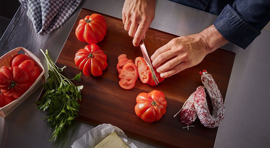 Person schneidet mit einem Messer der Classic-Serie von Wüsthof Tomaten auf Holzbrett, daneben Petersilie, Salami und Butter auf Edelstahloberfläche.
