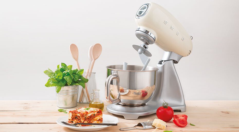 Küchenmaschine von SMEG mit Rührschüssel, neben Holzlöffeln, Pflanzen, Tomaten und einem tellergedeckten Stück Lasagne.