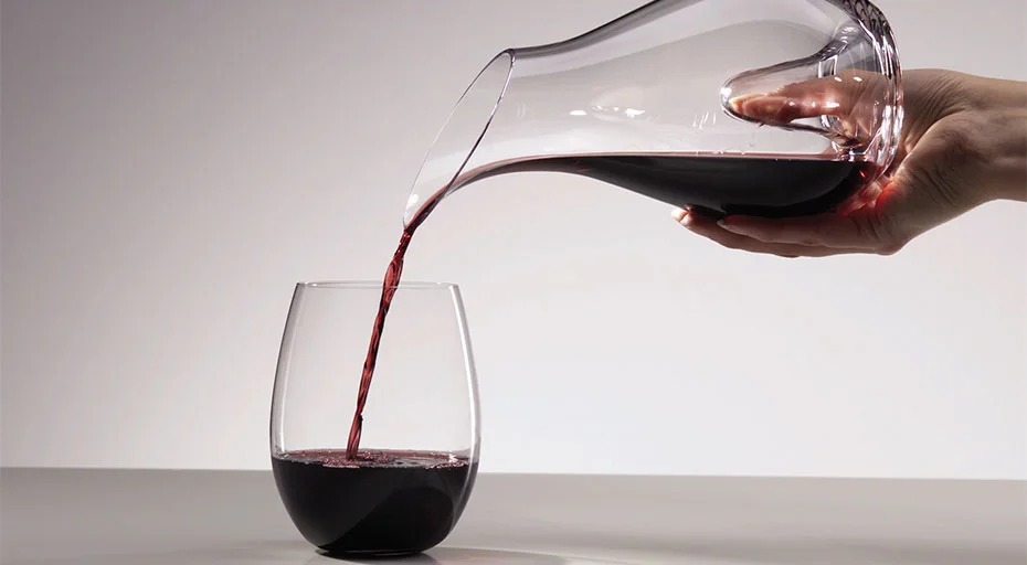 Eine Hand gießt Rotwein aus einer Karaffe in ein Weinglas ohne Stiel auf einer grauen Oberfläche.