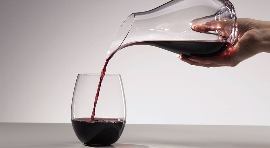 Eine Hand gießt Rotwein aus einer Karaffe in ein Weinglas ohne Stiel auf einer grauen Oberfläche.