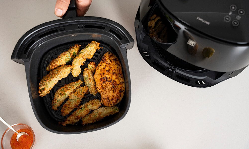 Luftfritteuse mit panierten, goldbraunen Avocado und einem Stück Hänchen. Daneben eine Schüssel mit roter Sauce.