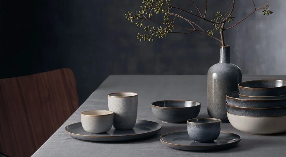 Keramikgeschirr der Serie Saisons von ASA auf Tisch: Tassen, Schalen und Teller in Grautönen; Vase mit Zweig, minimalistischer Stil.