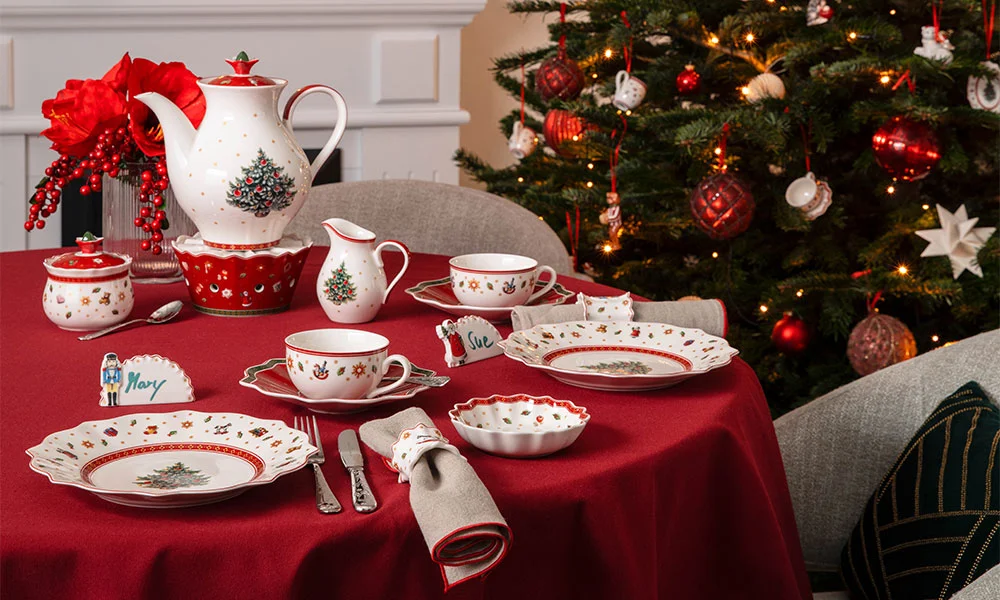 Villeroy &amp; Boch Toy's Delight Porzellan auf weihnachtlich gedecktem Tisch mit rotem Tischtuch, Geschirr mit Weihnachtsmotiven, Weihnachtsbaum im Hintergrund.